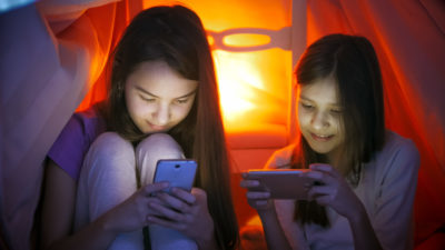 Portrait of two sisters in pajamas using smartphones under blanket at night