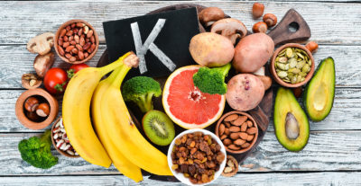 Foods containing natural potassium. K: Potatoes, mushrooms, banana, tomatoes, nuts, beans, broccoli, avocados. Top view. On a white wooden background.