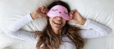 Above top view head shot happy millennial mixed race healthy woman lying in bed with sleeping mask, waking up after sweet dreams. Smiling young relaxed lady feeling energetic in morning at home.